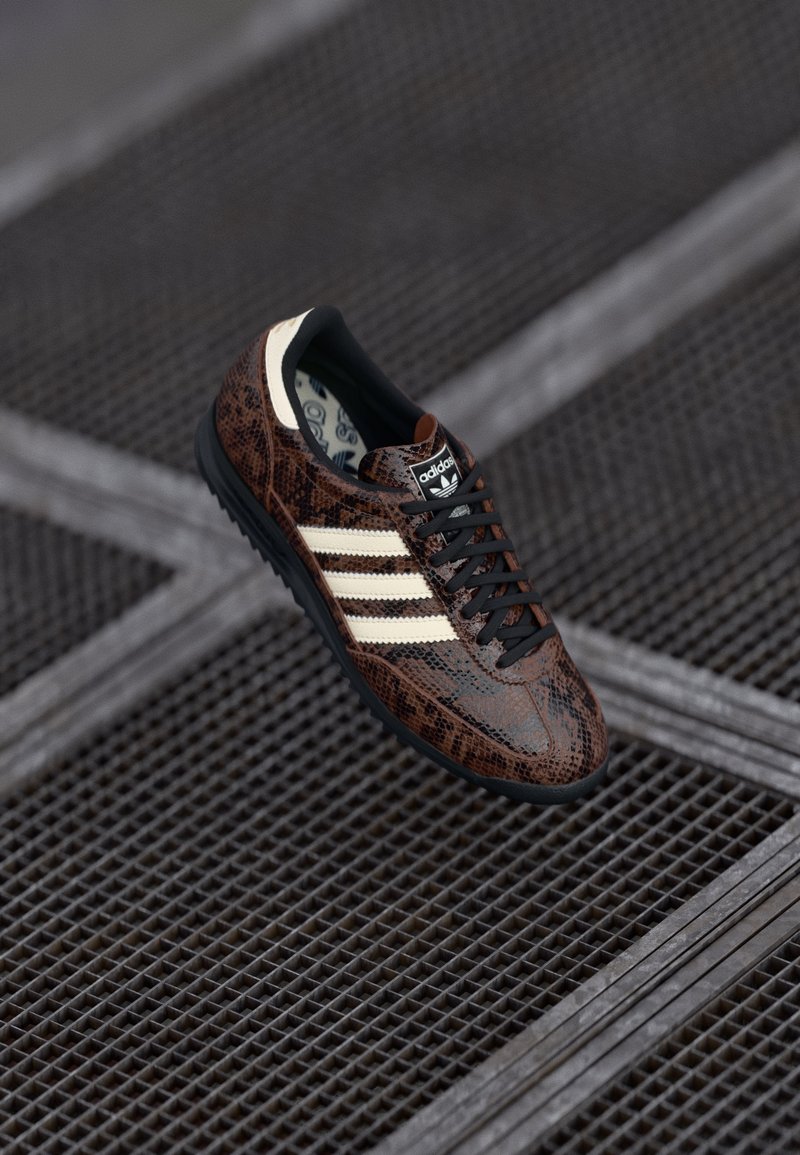 Sneaker a serpente marrone con lacci neri, strisce bianche e suola nera. Logo visibile sulla linguetta. Parte superiore in tessuto e parte inferiore in gomma.