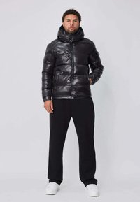 Veste matelassée noire avec un col montant et une fermeture éclair, présentant une texture brillante. Associée à un pantalon noir ample et des baskets blanches.