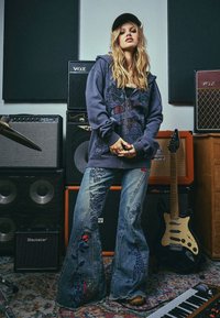 Oversize sinih džinsi s cvjetnim vezom, upareni s tamnosivim duksericom na zip s grafičkim uzorcima. Dodaci uključuju kapu i prstene.