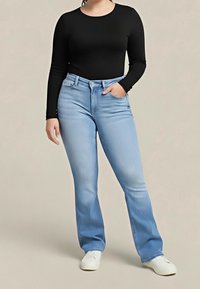 Personne portant un haut à manches longues noir, un jean bleu clair avec une coupe fuselée, et des baskets blanches, debout avec une main dans la poche.