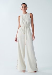 Svetlo bež kombinezon z zoštíhlenim v pasu, širokim krojem hlač in detajli z zavezovanjem na strani. Izdelan iz mehke, teksturirane tkanine.