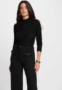 Donna con lunghi capelli scuri che indossa un top a maniche lunghe con stampa leopardata nera, pantaloni neri a vita alta con tasche frontali, orecchini a cerchio dorati e braccialetto dorato.