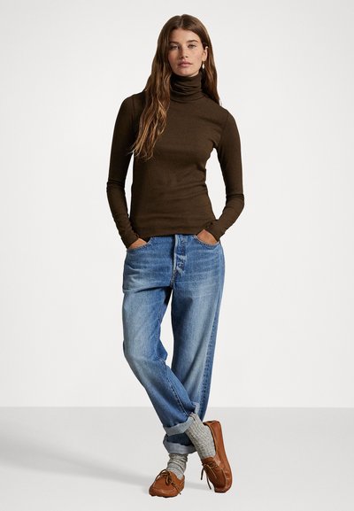 Brauner Rollkragenpullover, blaue hochtaillierte Jeans, graue Socken und braune Mokassins. Das Model posiert mit den Händen in den Taschen, lässiger Schnitt.