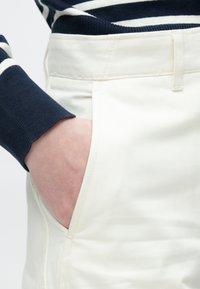 Kremfargede bukser med en glatt tekstur, med sidelommer og en tettsittende design. En marineblå genser med striper er synlig ved ermet.