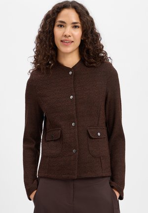 Vrouw met krullend donker haar, gekleed in een donkerbruine getextureerde jas met knopen en twee voorzakken met klep, handen in de zakken, tegen een witte achtergrond.