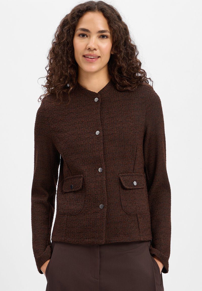 Woman with curly dark hair wearing a dark brown textured buttoned jacket with two front flap pockets, hands in pockets, on white background.