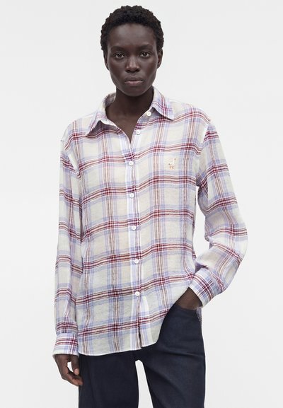 Γυναίκα που φοράει λευκό, κόκκινο και μπλε καρό πουκάμισο με κουμπιά και μακριά μανίκια, ένα χέρι στην τσέπη σκούρου παντελονιού, ουδέτερο φόντο.