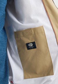 Tăsătură din plasă albă cu un buzunar orizontal maro, având o etichetă cu un logo albastru. Lângă aceasta se află un material texturat de culoare albastră.