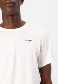 Bílý tričko G-Star RAW, kulatý výstřih, krátké rukávy, se malým černým logem na hrudi, vyrobeno z měkké bavlněné tkaniny, hladká textura.