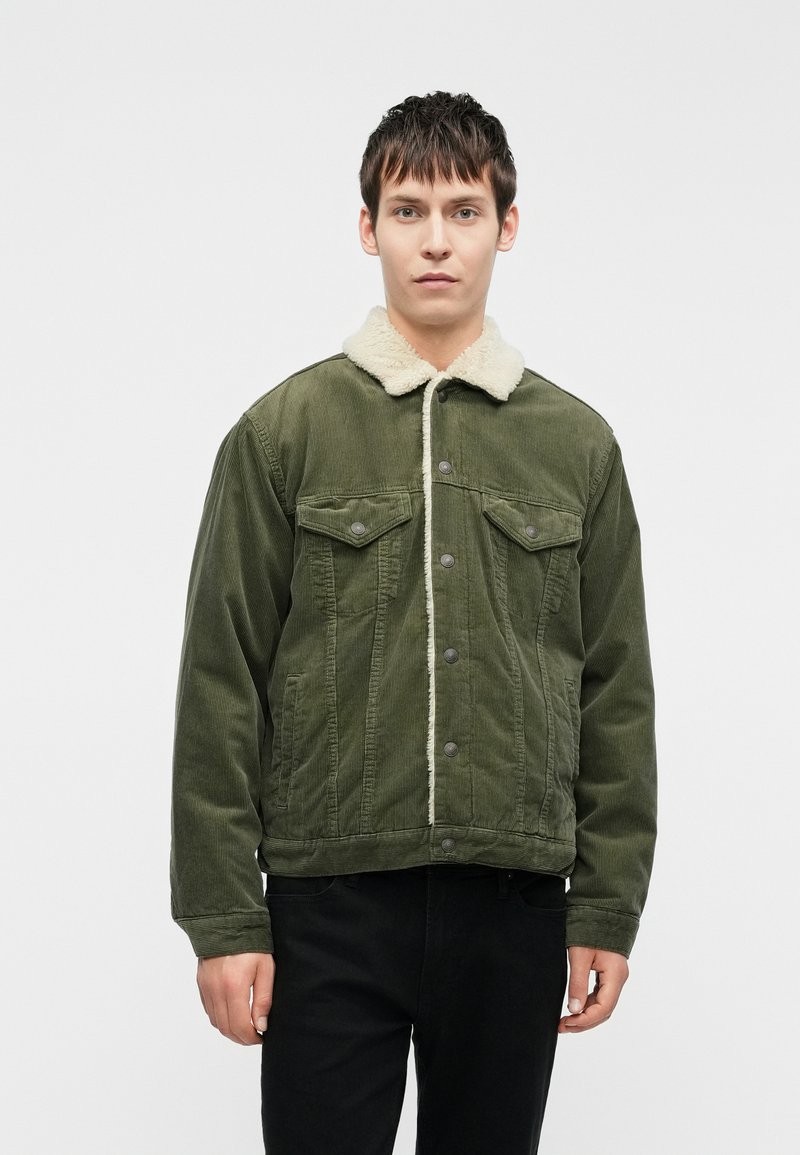 Casaco de veludo verde com um colarinho de pelo sintético creme, apresentando bolsos frontais e fecho de botão, tecido texturado e um corte descontraído.