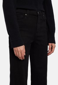 Jeans de mezclilla negra con corte recto, que presentan un cierre de botón y cremallera, cinco bolsillos y mangas de suéter de punto negro acanalado.