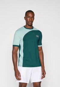 Camisa deportiva de manga corta con un diseño de bloques de color en azul verdoso y azul claro, tela texturizada y un logo en el pecho.