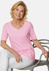 Helles rosa T-Shirt mit kurzen Ärmeln und V-Ausschnitt, verziert mit dekorativen Nieten entlang des Ausschnitts. Glatter Stoff mit lockerer Passform. Getragen zu weißen Hosen.