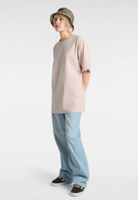 Hellbeige übergroßes T-Shirt mit gelben Grafiken, kombiniert mit hellblauen weit geschnittenen Jeans und einem Camouflage-Bucket-Hat. Schwarze Sneakers runden den Look ab.