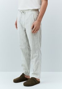 Pantalon gris clair texturé à rayures verticales, avec une taille élastique à cordon de serrage et des poches latérales. Porté avec des sabots foncés.