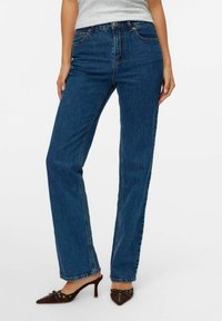 Woman wearing straight-leg blue denim jeans, gray top, and brown pointed-toe heeled shoes with small bows.