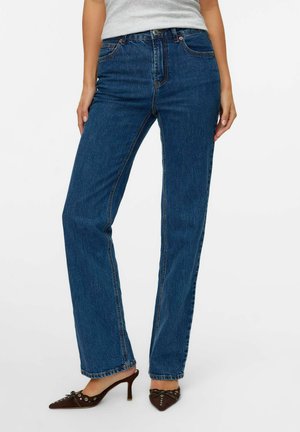 Vrouw in een rechte spijkerbroek in blauwe denim, grijs top en bruine puntige hakken met kleine strikjes.