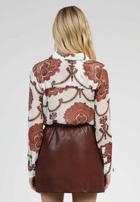 Blouse à manches longues avec un motif floral dans des tons chauds sur un fond blanc, associée à une mini-jupe en faux cuir marron.