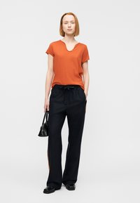 Κοντομάνικη μπλούζα σε χρώμα σκουριάς με στρογγυλή λαιμόκοψη, συνδυασμένη με μαύρο παντελόνι με φαρδιά πόδια και πορτοκαλί πλευρικές ρίγες. Αξεσουάρ περιλαμβάνουν μια μικρή μαύρη τσάντα.