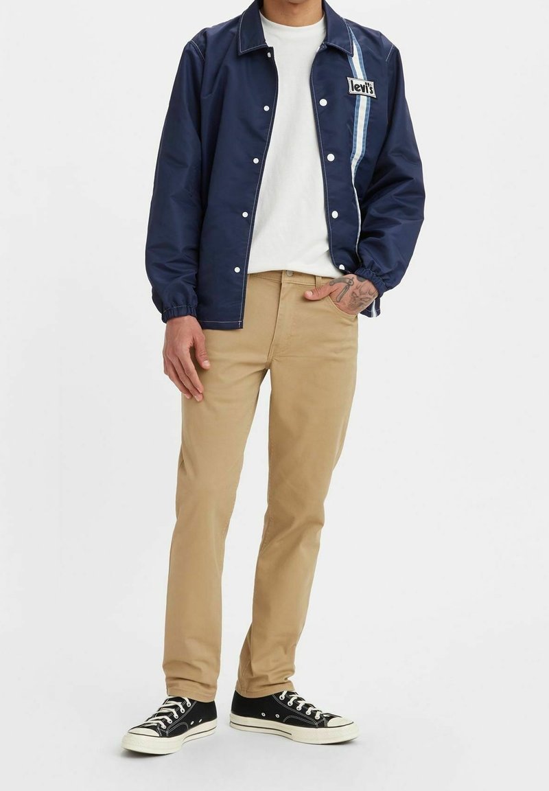 Hombre vestido con chaqueta navy de Levi's, camiseta blanca, pantalones beige y zapatillas negras, de pie con una mano en el bolsillo frente a un fondo blanco.