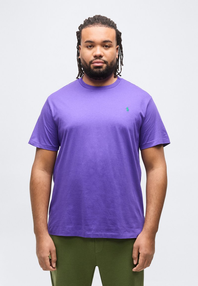 Άνδρας με πλεκτά μαλλιά και γένια, που φοράει μοβ μπλουζάκι με στρογγυλή λαιμόκοψη και παντελόνι ελιάς, στέκεται μπροστά σε απλό φωτεινό φόντο.