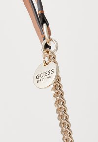 Kultaketju, jossa on ympyrätunnus kaiverrettuna "GUESS EST.1981" ja joka on kiinnitetty mustaan ja ruskeaan nahkahihnaan.