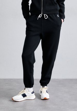 Black cotton joggers with a cuffed hem and white drawstring. Paired with white sneakers featuring navy accents and a textured sole.