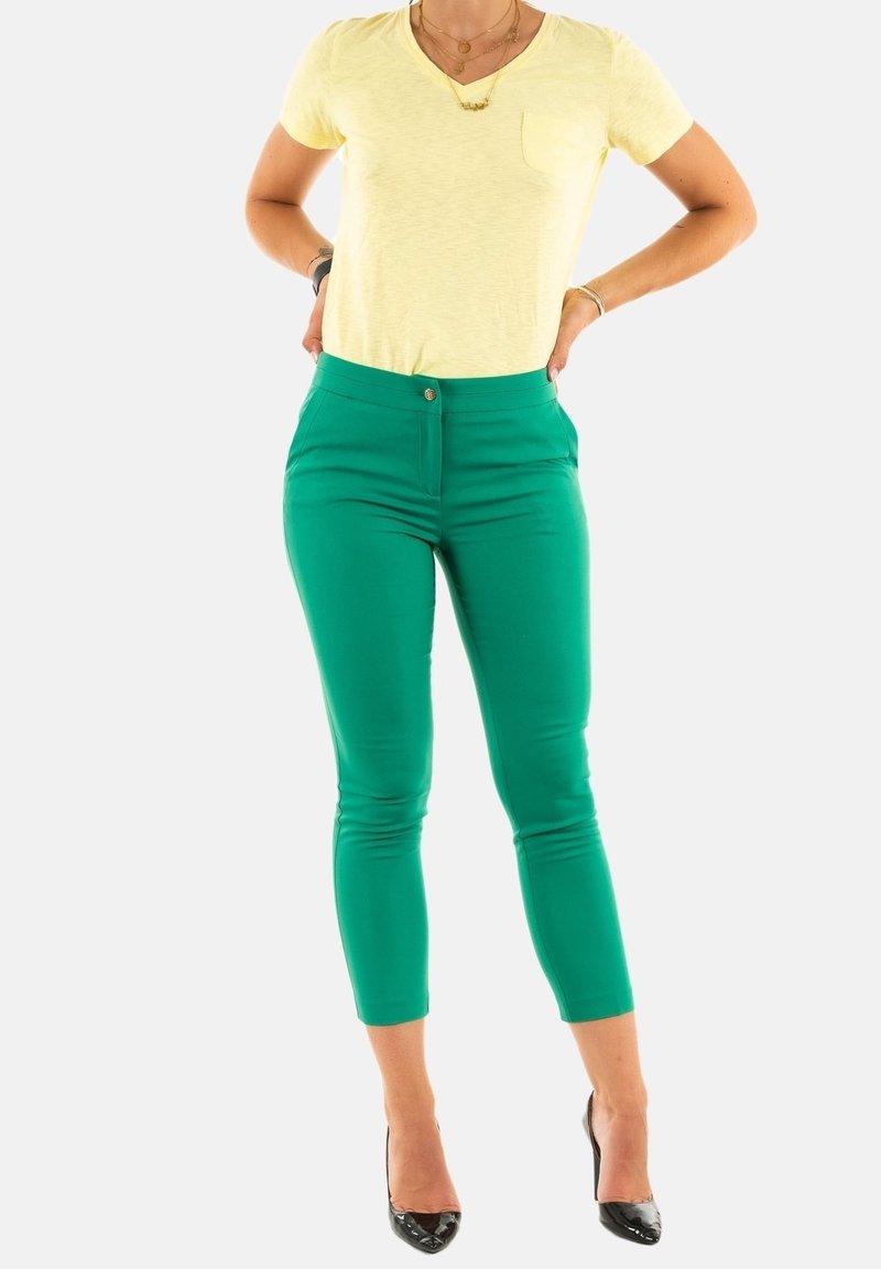 Personne portant un pantalon slim vert, une chemise jaune à manches courtes, des talons noirs et des colliers en or, debout les mains sur les hanches.