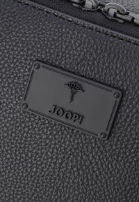 Sort tekstureret materiale med en forhøjet logoplade. Designet har subtile mønstre og metal detaljer, der understreger et slankt, moderne udseende.