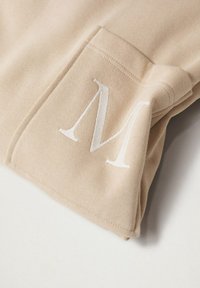 Beige Sweatshirt mit einer Vordertasche, die mit einem großen weißen gestickten "M" verziert ist, aus weichem, strukturiertem Stoff gefertigt.