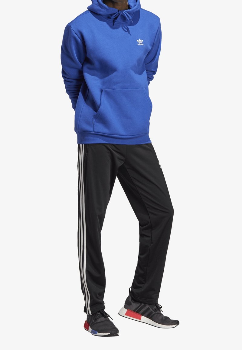 Sininen huppari, jossa on nyöri ja etutasku, yhdistetty mustiin urheiluhousuihin, joissa on kolme valkoista raita. Mustat lenkkarit punaisilla yksityiskohdilla.