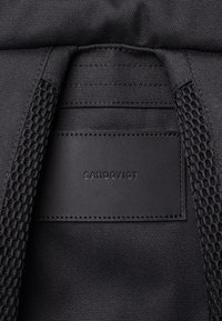 Gros plan sur un tissu noir avec un patch en cuir cousu affichant le nom de la marque Sandqvist sur un sac à dos noir texturé.