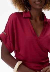 Rode satijnen blouse met kraag, korte mouwen en een wikkelontwerp. Bevat een label op de mouw en een oversized crèmekleurige armband om de pols.