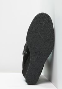 Chaussure en suede noire avec une semelle en caoutchouc texturé. Présente un détail de sangle et un logo de la marque embossé. Vue depuis la base sur un fond clair.