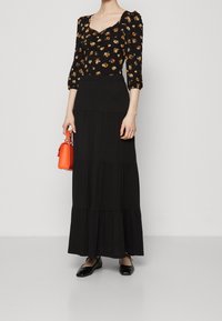 Kvinna iklädd en svart blommig topp med puffärmar, en lång svart lagerkjol, svarta ballerinas och håller en liten orange handväska.