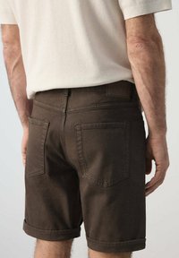 Hombre con pantalones cortos de dobladillo enrollado de color marrón oscuro y una camisa de manga corta beige clara, visto desde atrás a nivel de la cintura.