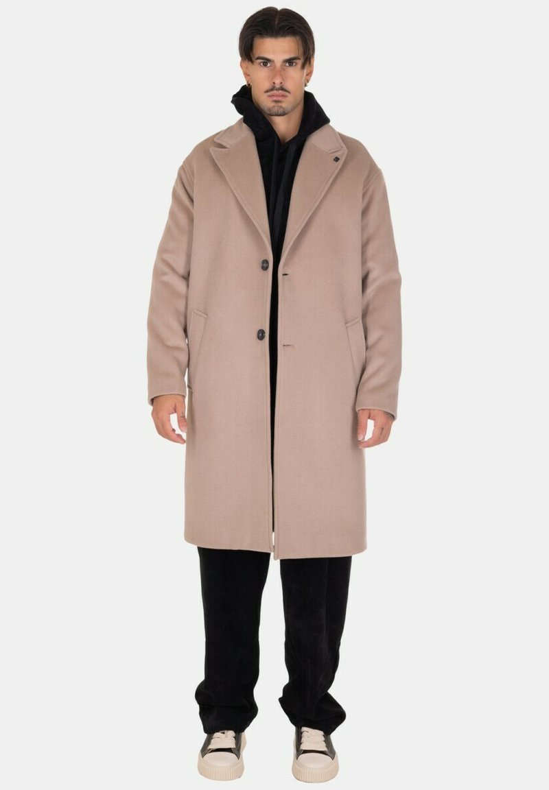 Cappotto di lana beige con collo a punta, chiusura a un bottone, tasche laterali, indossato sopra un hoodie nero, abbinato a pantaloni neri e sneakers.