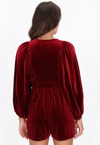 Mono corto de terciopelo rojo con mangas largas abullonadas, cintura elástica y detalles fruncidos en la parte trasera. Textura suave y diseño de shorts cortos.