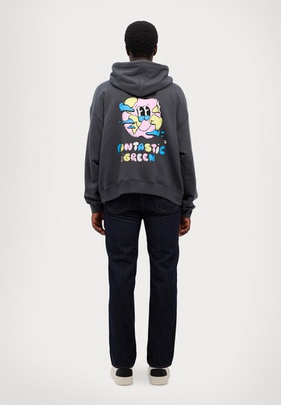 Grå hættetrøje med et farverigt graphic på ryggen, med teksten "FANTASTISK GRØN" under et legende design, parret med mørke jeans og sneakers.