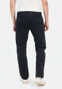 Zwarte corduroybroek met rechte pijpen, voorzien van twee achterzakken en een subtiele textuur. Draagt witte sneakers.