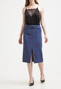 Femme portant une camisole noire ornée de dentelle, une jupe midi bleue ceinturée avec une fente devant et des poches plaquées, ainsi que des chaussures à talons hauts noires.