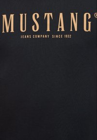 Tricou negru din bumbac cu inscripție în text auriu care citește "MUSTANG JEANS COMPANY ÎNCA DIN 1932." Textură netedă și design clasic cu guler rotund.