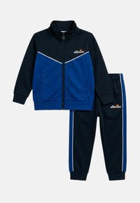 Veste zippée bleu marine et bleue avec passepoil blanc et pantalon assorti bleu marine avec des bandes latérales bleues, tous deux arborant le logo ellesse.