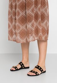Black slide sandals with metallic gold embellishments, flat sole, and wide straps. Worn with a brown patterned midi dress.