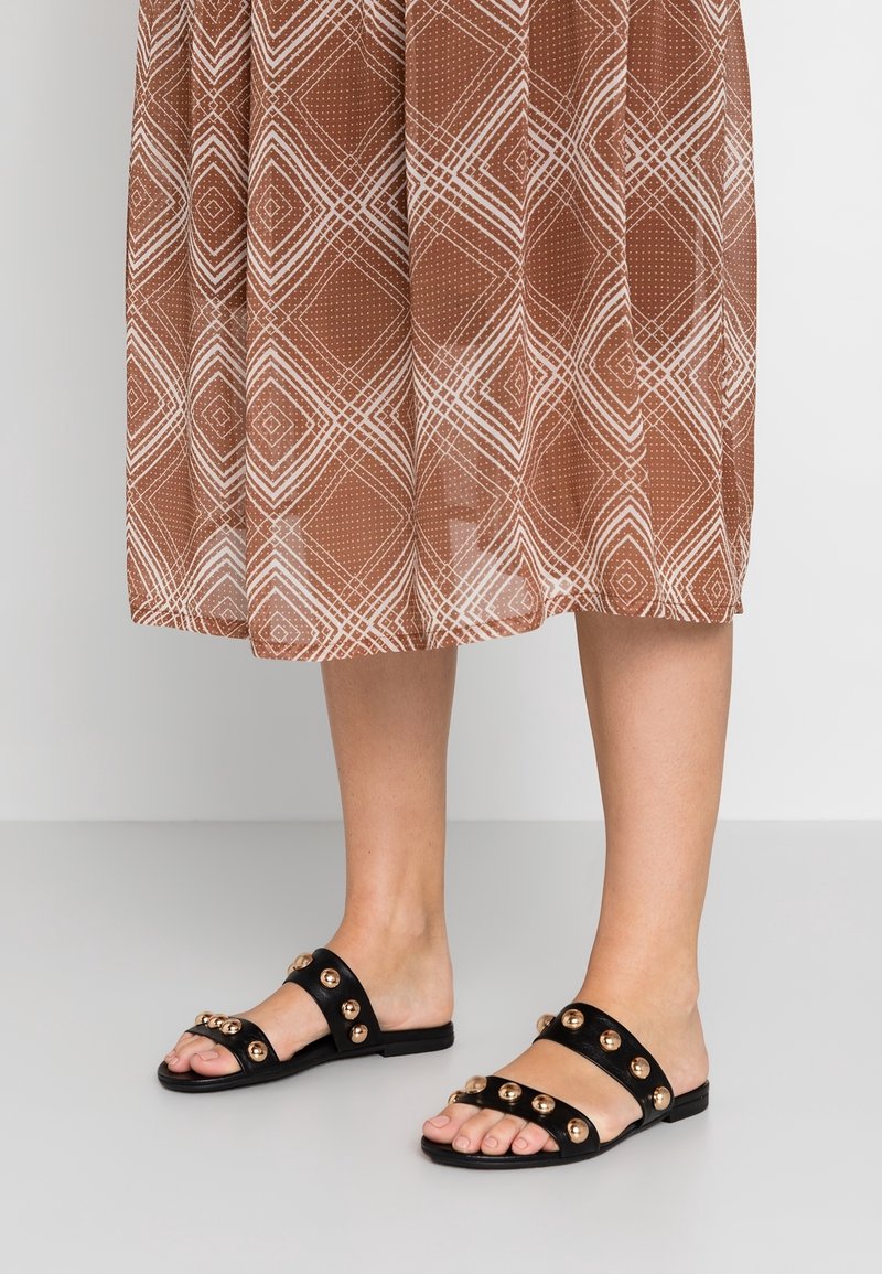 Black slide sandals with metallic gold embellishments, flat sole, and wide straps. Worn with a brown patterned midi dress.