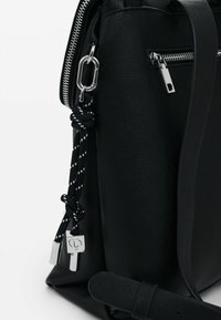 Schwarze, strukturierte Ledertasche mit silbernen Beschlägen, darunter ein Reißverschluss und dekorative Akzente aus schwarz-weißen geflochtenen Kordeln.