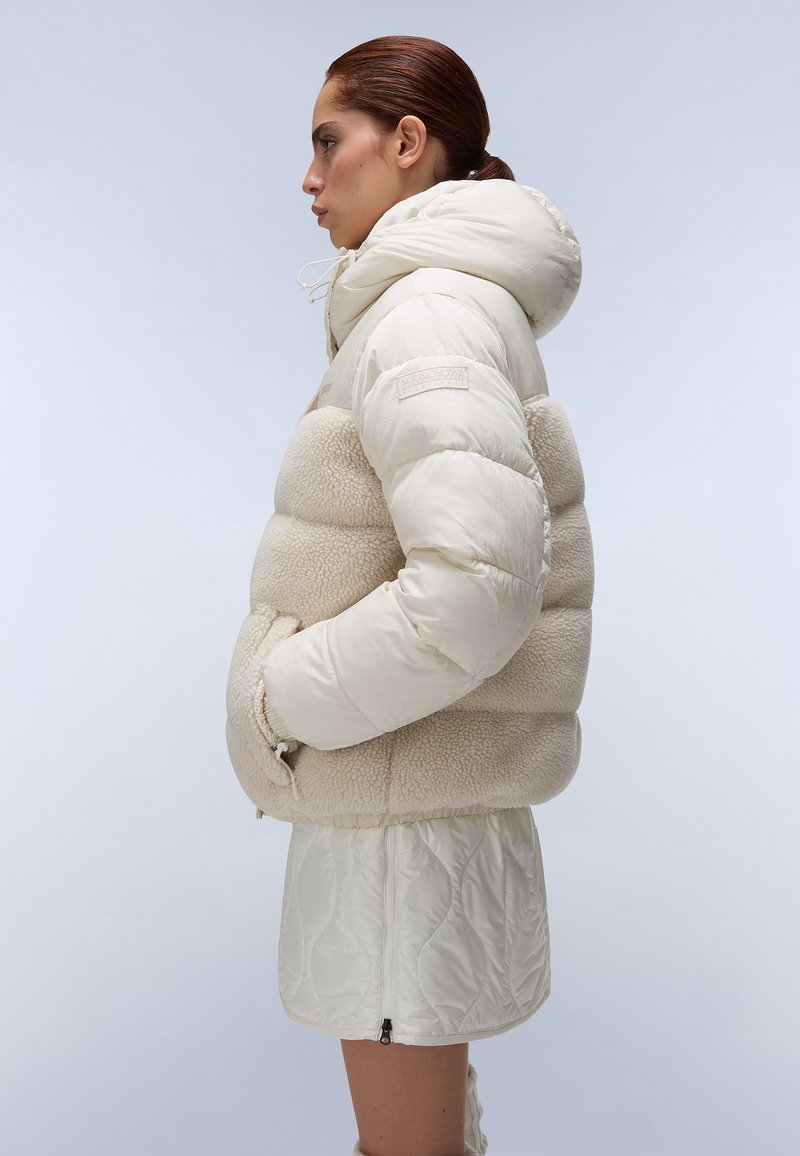 Donna di profilo che indossa una giacca a vento imbottita con cappuccio di colore crema, con una sezione centrale in pile e una gonna trapuntata coordinata, su uno sfondo chiaro.