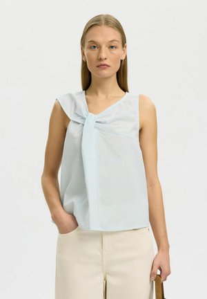 Femme aux cheveux blonds raides portant un chemisier sans manches bleu clair noué devant et un pantalon crème, debout sur un fond clair uni.