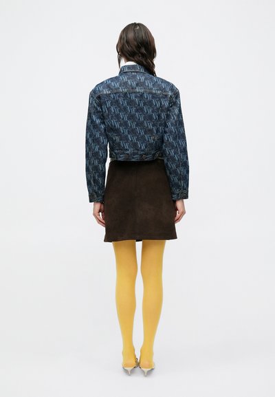 Džínová bunda s modrým logom, hnedá semišová sukňa, žlté pančuchy a strieborné podpätky, zobrazené zozadu na jednoduchom pozadí.