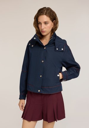 Jonge vrouw met kort bruin haar, gekleed in een marineblauwe jas met zilveren knopen en een bordeaux plissérok, staand voor een effen achtergrond.